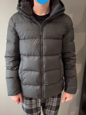 Svart J.Lindeberg jacka - Snygg svart pufferjacka från J.Lindeberg med huva och dragkedja framtill. Jackan har en quiltad design med breda kanaler och diskret logga på bröstet. Perfekt för kalla vinterdagar och riktigt skön att bära tack vare det vadderade materialet.