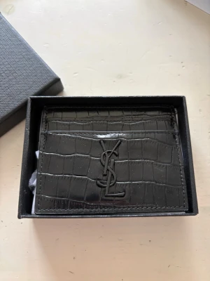 Svart Saint Laurent kortfodral i läder - Snyggt svart kortfodral från Saint Laurent i präglat läder med krokodilstruktur. Fodralet har flera kortfack och en stilren YSL-logga framtill i svart metall. Perfekt för dig som vill ha en exklusiv och trendig accessoar i fickan.