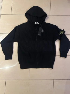 Svart stickad cardigan från Stone Island - Säljer en svart stickad cardigan från Stone Island med dragkedja framtill och ikonisk patch på ärmen. Jackan har ribbad struktur, huva och långa ärmar. Perfekt för dig som vill ha en clean och stilren look med streetwear-vibe.