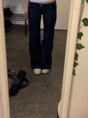 Svarta bootcut jeans med låg midja - Säljer ett par svarta bootcut jeans med låg midja och klassisk femficksmodell. Jeansen har utsvängda ben och silverfärgade knappar. Materialet är denim med stretch för skön passform. Perfekta för dig som gillar en retroinspirerad look.
