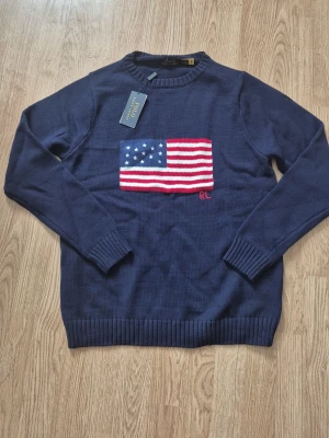 Marinblå stickad tröja Polo Ralph Lauren - Stickad marinblå tröja från Polo Ralph Lauren med amerikansk flagga broderad på bröstet och RL-detalj. Tröjan har rund halsringning, ribbade muddar och är gjord i mjukt material. Perfekt för dig som gillar klassisk och stilren design.