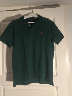 Mörkgrön pikétröja från Ralph Lauren - Klassisk mörkgrön pikétröja från Ralph Lauren med korta ärmar och krage. Tröjan har den ikoniska röda logotypen broderad på bröstet och stängs med två knappar vid halsen. Tillverkad i mjuk bomull för en bekväm känsla.