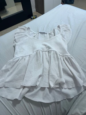 Vit volangblus från 157 - Supersöt vit blus från 157 med volanger och lös passform. Blusen har korta ärmar med volangdetaljer och är tillverkad i ett lätt, strukturerat tyg som känns luftigt och fräscht. Perfekt för dig som gillar en romantisk och trendig stil.