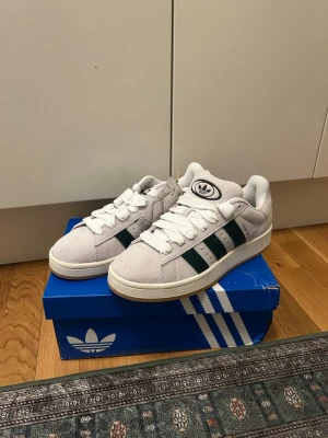 Helt nya Adidas Campus vita/gröna sneakers - Säljer ett par helt nya Adidas Campus sneakers i vitt mockaskinn med mörkgröna klassiska ränder på sidorna. Skorna har vit sula, vita skosnören och Adidas-logga på plösen och hälen. Stilren och tidlös modell som passar till det mesta. Lådan är lite trasig.