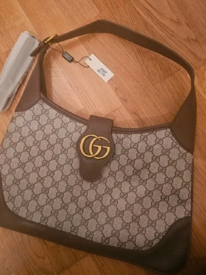  GG Supreme axelremsväska brun - Snygg axelremsväska i beige och brunt GG Supreme-mönster. Väskan har detaljer i brunt läder, guldigt GG-spänne framtill och bred axelrem. Klassisk halvmåneformad modell med exklusiv känsla och tydlig logga.