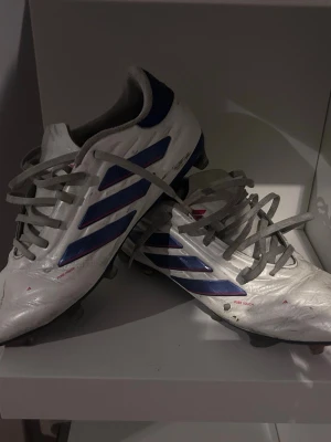 Adidas Copa vita fotbollsskor - Säljer ett par vita Adidas Copa fotbollsskor med blå detaljer och grå snörning. Skorna har klassiska tre ränder på sidan och är tillverkade i syntetmaterial med FG-dobbar för spel på naturgräs. Perfekta för dig som vill ha bra touch och kontroll på planen.