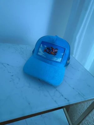 Blå Superman keps med mesh - Snygg blå keps med Superman-tryck framtill och mesh på sidorna för extra ventilation. På sidan finns ett vitt 'W'-märke broderat. Justerbar baktill för perfekt passform. Perfekt för dig som gillar superhjältar och streetwear.