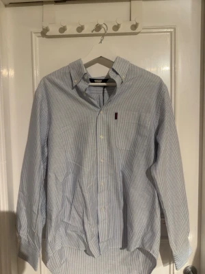 Randig blåvit skjorta från Sebago - Klassisk blåvit randig skjorta från Sebago  med lång ärm och knappar framtill. Skjortan har en bröstficka med diskret logga och är tillverkad i mjuk bomull. Perfekt för en clean och stilren look.