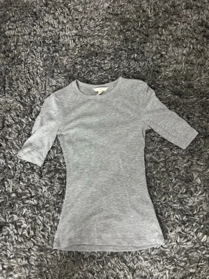 Ljusgrå ribbad topp med halvlång ärm - Snygg ljusgrå ribbad topp med rund hals och halvlånga ärmar. Figurnära passform som ger en clean och stilren look. Perfekt att matcha med jeans eller kjol för en enkel men trendig outfit.