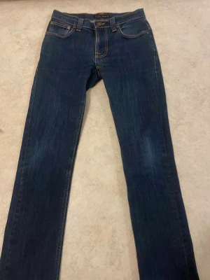 Mörkblå jeans från Nudie Jeans - Säljer ett par mörkblå jeans från Nudie Jeans med klassisk femficksmodell och raka ben. Jeansen har kontrastsömmar och snygga broderade detaljer på bakfickorna. Tillverkade i slitstark denim, perfekta för en avslappnad stil.