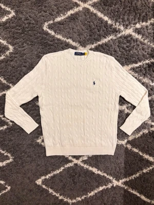 Vit Polo Ralph Lauren tröja  - Kabelstickad vit tröja från Polo Ralph Lauren med rund halsringning och broderad marinblå logga på bröstet. Tröjan har långa ärmar och ribbade muddar vid ärmslut och nederkant. Jag är 180cm och väger 180kg och den passar mig perfekt.