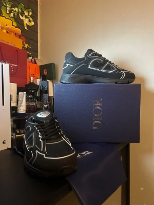 Svarta Dior B30 sneakers - Snygga svarta Dior B30 sneakers med vita detaljer och reflekterande linjer. Skorna har en chunky sula, mesh och mocka-paneler samt CD-logga på hälen. Perfekta för dig som gillar exklusiva och trendiga sneakers med sportig vibe.