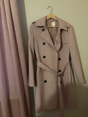 Ljusrosa trenchcoat med knytskärp - Stilren ljusrosa trenchcoat med dubbelknäppning och svarta knappar. Knytskärp i midjan och klassisk krage. Långa ärmar och två fickor framtill. Perfekt för vår och höst. 