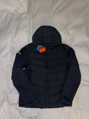 Superdry cardigan hoodie mörkblå - Snygg svart hoodie-cardigan från Superdry med vadderad framsida och stickade mörkgrå ärmar. Jackan har dragkedja, huva och Superdry-logga på ärmen. Perfekt för lager-på-lager och streetwear-stil. Självklart äkta och pris kan diskuteras