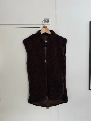 Mörkbrun teddyväst med dragkedja - Mysig mörkbrun teddyväst med hög krage och hel dragkedja framtill. Västen har två fickor med dragkedja och är tillverkad i ett fluffigt, mjukt material. Perfekt för lager på lager och ger en avslappnad, trendig look.