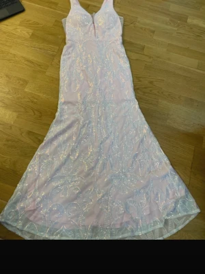 Lång rosa paljettklänning med djup v-ringning - Superfin långklänning i ljusrosa med glittriga paljettdetaljer och djup v-ringning. Klänningen har ett elegant mönster och är ärmlös, perfekt för bal eller fest. Materialet känns lyxigt och skimrar snyggt i ljuset.