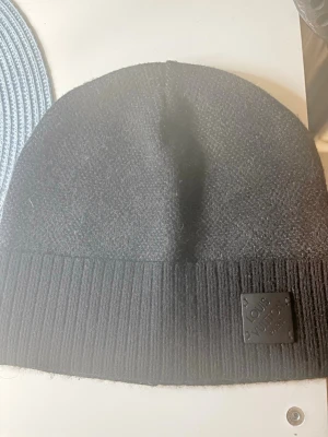 Svart mössa från L - Snygg svart mössa från L med diskret logopatch framtill. Mössan har ribbad kant och är tillverkad i ett mjukt, stickat material som håller dig varm. Perfekt accessoar för kalla dagar och en stilren look.