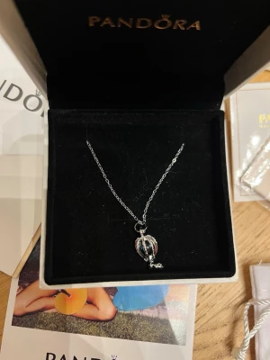 Pandora silverhalsband med pärlor - Säljer ett elegant silverhalsband från Pandora med ett hängsmycke dekorerat med små vita pärlor. Kedjan är tunn och smidig, och halsbandet levereras med originalförpackning, påse och certifikat. Perfekt accessoar för dig som gillar stilrena smycken.