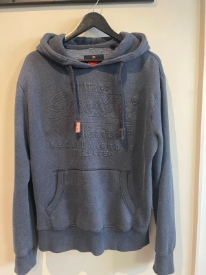 Mörkblå hoodie från Superdry - Snygg mörkblå hoodie från Superdry med broderad logga på bröstet och klassisk känguruficka. Hoodien har dragsko i huvan och ribbade muddar vid ärmar och nederkant. Perfekt för en avslappnad och trendig stil. Pris kan diskuteras 