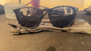 Svarta Ray-Ban solglasögon - Snygga svarta solglasögon från Ray-Ban med klassisk rundad form och mörka glas. Perfekta för att ge en cool vibe till din outfit. Kommer med en praktisk tygpåse för förvaring.