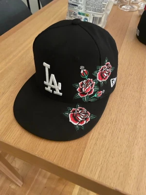 Svart LA-keps med rosbrodyr - Säljer en svart New Era-keps med LA-logga framtill i vitt och snygga broderade röda rosor med gröna blad på sidan och skärmen. Klassisk flat brim-modell och MLB-logga bak. Perfekt för dig som vill sticka ut med en unik accessoar.