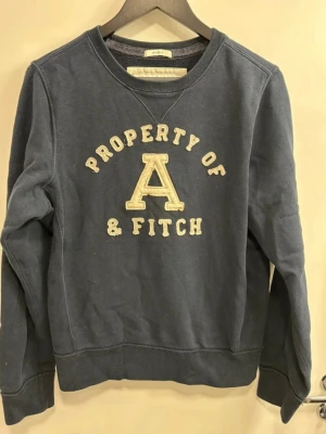 Blå sweatshirt från Abercrombie & Fitch - Mörkblå sweatshirt från Abercrombie & Fitch med broderad text 'Property of A & Fitch' på bröstet. Klassisk rund halsringning och ribbade muddar vid ärmslut och nederkant. Perfekt för en avslappnad och cool stil.