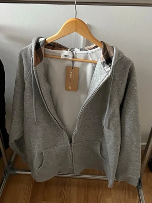 Grå zip-hoodie från Burberry - Snygg grå hoodie med dragkedja från Burberry. Klassisk huva med justerbara snören och ikoniskt rutigt foder i huvan. Mjukt material och två fickor framtill. Perfekt för en avslappnad och trendig stil.