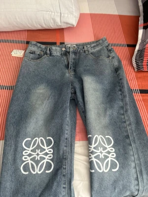 Blå baggy jeans från Loewe - Säljer ett par blå baggy jeans från Loewe med vita grafiska tryck på båda benen. Jeansen har klassisk femficksmodell, knappgylf och en grön patch med Loewe-logga bak. Tillverkade i denim med en cool tvättad look och bred passform. HELT NY O OANVÄND, 1-1