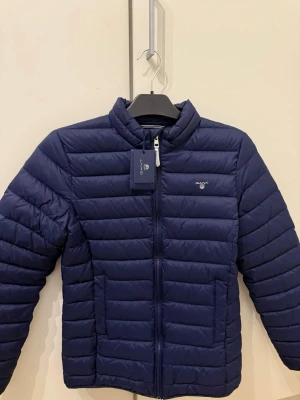 Mörkblå pufferjacka från GANT - Snygg mörkblå pufferjacka från GANT med logga på bröstet. Jackan har horisontella sömmar, dragkedja framtill och två sidofickor. Nyskick, aldrig använd! Storlek 176