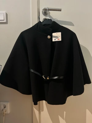 Svart cape-kappa med bälte - Stilren svart cape från Hk & City med elegant knäppning vid halsen och ett smalt bälte i midjan. Kappan har en klassisk, vid siluett som ger en lyxig känsla. Perfekt för dig som vill ha en chic och tidlös look. Tillverkad i Italien 