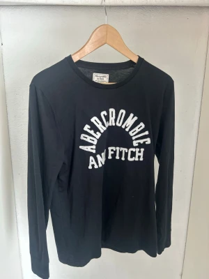 Svart långärmad topp Abercrombie - Svart långärmad topp från Abercrombie & Fitch med vit text framtill. Klassisk rund halsringning och normal passform. Tillverkad i mjuk bomull som känns skön mot huden. Perfekt för en avslappnad och stilren look.
