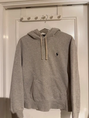Grå hoodie från Ralph Lauren - Snygg grå hoodie från Ralph Lauren med klassisk huva och dragsnören. Har en broderad logga på bröstet och en stor känguruficka framtill. Perfekt för en chill och avslappnad stil. Mjuk bomullsblandning som känns skön mot huden.