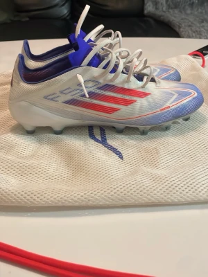 Adidas f50 elite ag fotbollsskor - Snygga Adidas f50 ag elite fotbollsskor i vitt med röda och blå detaljer. Skorna har låg vikt, snörning och AG-dobbar för spel på konstgräs. Perfekta för dig som vill ha fart på planen. Använda max 3 gånger på konstgräs. Nypris ca 2-3 tusen