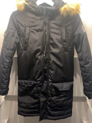 Vinson Polo Club med pälsluva mycket fint skick - Säljer en varm och stilren vinterjacka från Vinson Polo Club. Lång modell med huva och pälsdetalj, perfekt för kalla dagar.  Storlek: 12 (motsvarar ca S/M – se passform) Skick: mycket fint, sparsamt använd Färg: svart