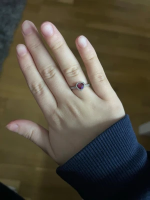 Silverring med röd hjärtformad sten - Fin silverfärgad ring med en hjärtformad röd sten i mitten. Ringen har små klara stenar runt hjärtat och längs bandet, vilket ger extra glans och en romantisk vibe. Perfekt accessoar för dig som gillar detaljer och vill ha något som sticker ut.