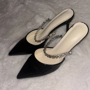 Svarta pumps med strassdetaljer - Säljer ett par eleganta svarta pumps med spetsig tå och hög klack. Skorna har en dekorativ rem med glittrande strass som ger extra lyxig känsla. Ovandelen är i blankt material och innersulan är ljus. Perfekta för dig som vill sticka ut.