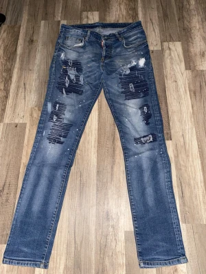blåa D2 slitna jeans med färgstänk - Ett par blåa D2 jeans med slitningar och hål på benen samt vita färgstänk för en cool, trendig look. Jeansen har fem fickor och en något tvättad finish. Perfekta för dig som gillar streetstyle och vill sticka ut.