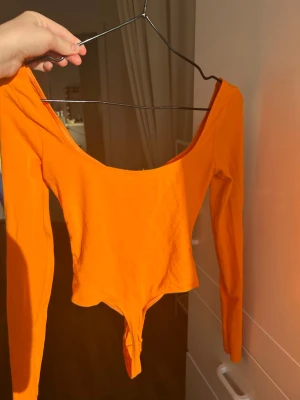 Orange långärmad body med knyt - Snygg orange body med långa ärmar och djup urringning. Framtill finns en knytning som ger en extra detalj. Tillverkad i stretchigt tyg som sitter tajt och bekvämt. Perfekt för dig som vill sticka ut med en färgstark look.