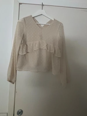 Beige blus med volang och prickar - Skriv om ni vill ha fler bilder!❤️Säljer en beige blus med långärmad design och diskreta prickar. Blusen har en volang framtill och är tillverkad i ett tunt, transparent material som ger en luftig känsla. Perfekt för att skapa en söt och trendig look.