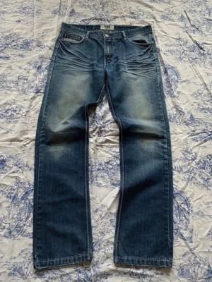 Blå raka jeans från NNB - Snygga blå jeans från NNB med raka ben och slitna detaljer framtill och baktill. Jeansen har klassisk femficksdesign, ljusare partier på lår och knän samt coola sömmar på baksidan av benen. Perfekta för en avslappnad streetstil.