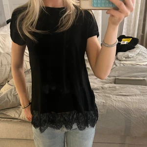 Svart topp med spetskant - Svart kortärmad topp med rund halsringning och snygg spetsdetalj längst ner. Toppens material känns mjukt och lätt, perfekt att styla med jeans eller kjol. Enkel men ändå med en extra touch tack vare spetsen.