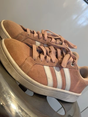 Rosa Adidas sneakers i mocka - Säljer ett par snygga rosa Adidas sneakers i mocka med klassiska vita ränder på sidorna. Skorna har matchande rosa skosnören och vit sula. Perfekta för dig som gillar en sportig och trendig stil.