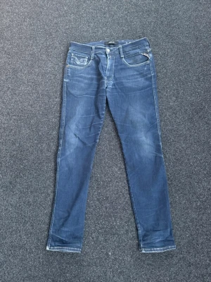 Blå slim jeans från Replay - Snygga blå slim jeans från Replay med klassisk femficksdesign och diskreta slitningar. Jeansen har låg midja, dragkedjegylf och är tillverkade i stretchigt denim för en bekväm passform. Perfekta för en avslappnad och trendig look.                                Storlek 32-32