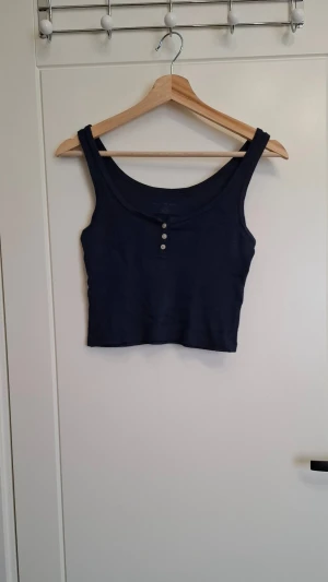 Mörkblå ribbad croppad linnetopp - Säljer en mörkblå croppad linnetopp från Brandy Melville. Toppen är ribbad och har tre dekorativa knappar framtill. Perfekt till jeans eller kjol för en enkel och snygg look.