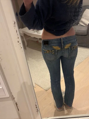 Blå bootcut jeans med detaljer - Säljer ett par blå bootcut jeans med snygga guldiga nitar och broderade vingar på bakfickorna. Jeansen har låg midja och klassisk femficksdesign. Perfekta för dig som vill sticka ut med coola detaljer och en skön passform.