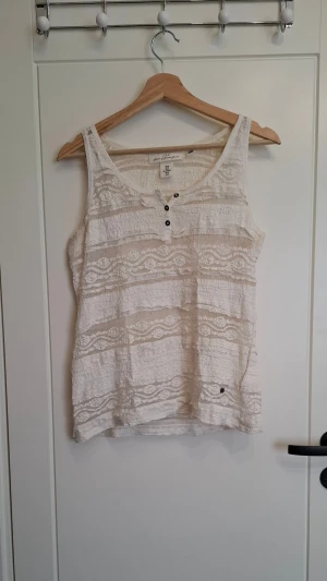Vit linnetopp från H&M L.O.G.G. - Säljer en vit linnetopp från H&M L.O.G.G. med dekorativa knappar framtill och spetsdetaljer i tyget. Toppen är ärmlös och har ett lätt strukturerat material med fina mönster i tyget. Perfekt för varma dagar och enkel att styla.