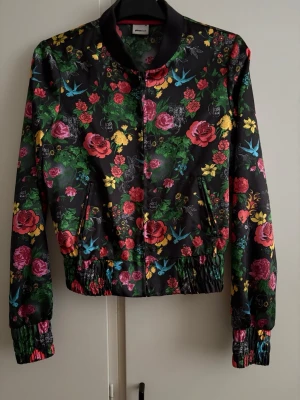 Blommig bomberjacka med fågelmotiv - Färgstark bomberjacka f med svart bas, täckt av blommor i rött, rosa, gult och grönt samt blå fåglar. Jackan har dragkedja, ribbade muddar och elastisk midja. Fickor på sidorna 