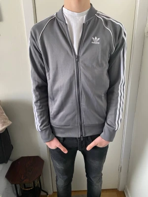 Grå Adidas ziptröja med vita ränder - Säljer en grå ziptröja från Adidas med klassiska vita ränder längs ärmarna och Adidas-logga på bröstet. Tröjan har hög krage, muddar vid ärmslut och nederkant samt fickor på sidorna. Perfekt för en sportig och avslappnad stil. Knappt använd alls i otroligt bra skick! 