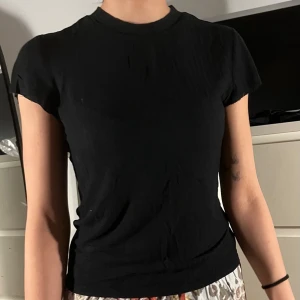 Svart tight t-shirt med rund hals - En enkel svart t-shirt med rund hals och korta ärmar. Modellen är tight och passar perfekt till jeans eller kjol. T-shirten är stilren och lätt att matcha med andra plagg.