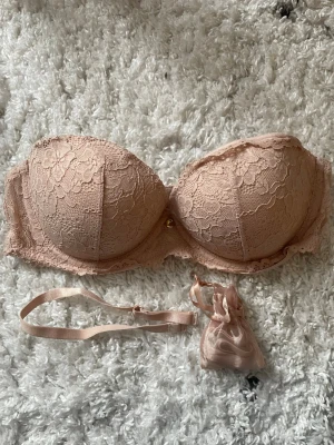 Ljusrosa/ beige spets-bh med guld detalj axelbandslös ca 75D (klädlapp saknas)  - Säljer en fin beige bh med vacker spets. - avtagbara axelband tillkommer   - vadderade kupor   Storlekslappen har tyvärr klippts bort. Jag har mätt den noggrant och omkretsen är ca 72 cm (väldigt stretchigt band) vilket motsvarar storlek 75  Kupa: rymlig och djup modell (ca 19 cm tvärs över insidan och 13 cm på höjden) uppskattas till en D-kupa.  Passar dig som har en smalare rygg, men behöver en djupare kupa än standard.   Passar perfekt under toppar med bara axlar. 
